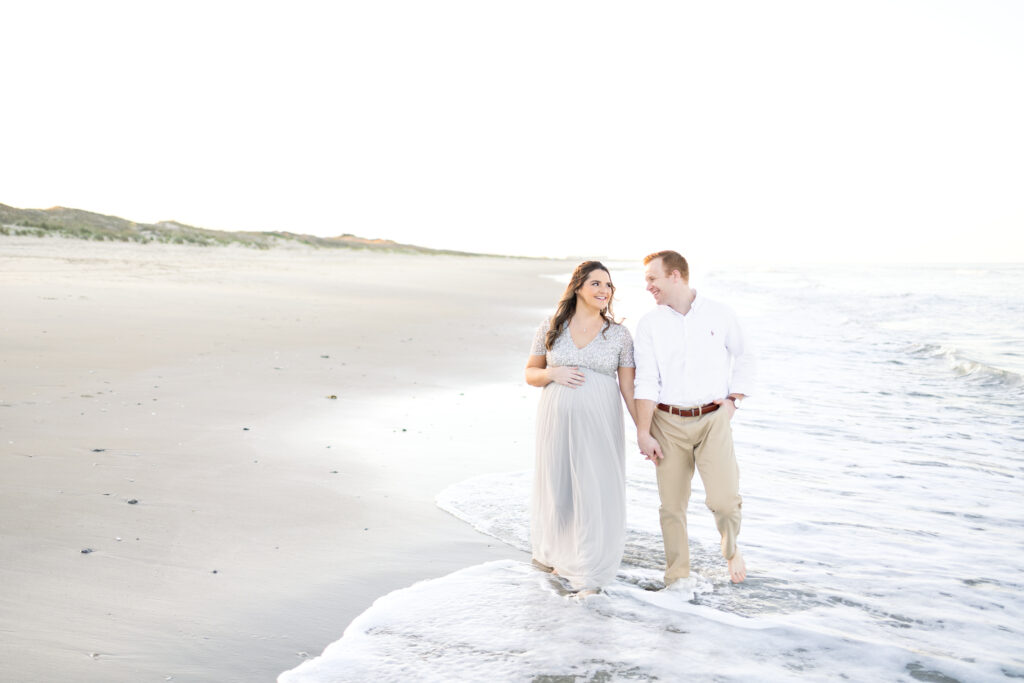 couple smiling and walking together during virginia beach maternity portraits with chelsey kraus photography 