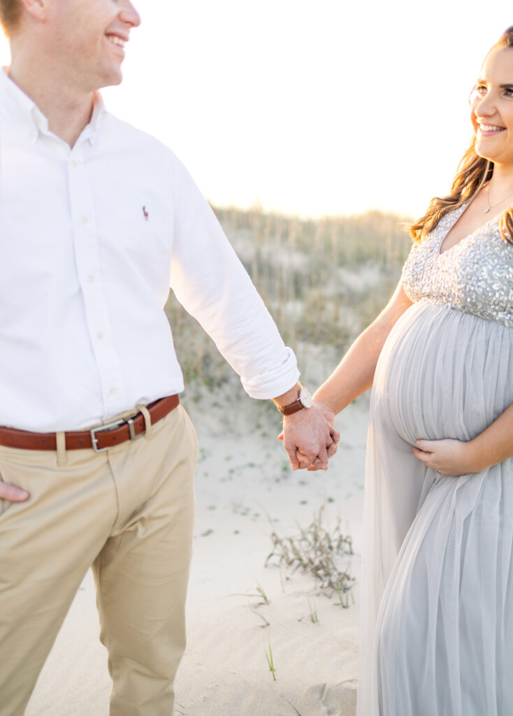 mom and dad holding hands and smiling at each other during maternity portraits in virginia beach