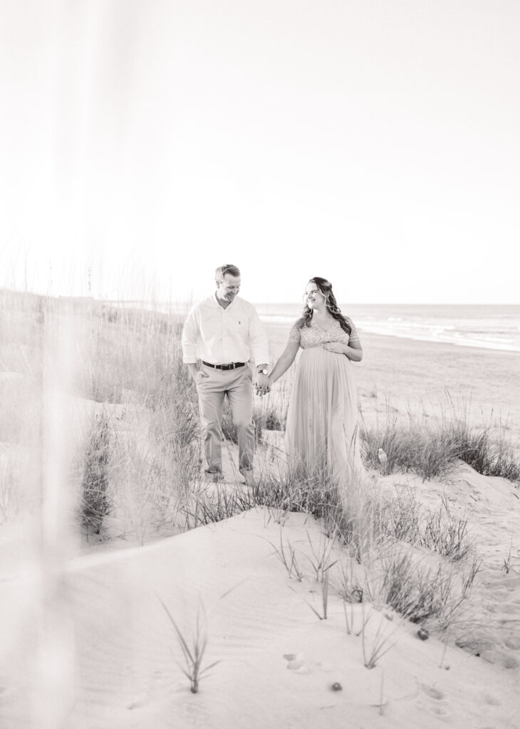 mom and dad to be walking and holding hands on the beach in virginia beach during maternity photos