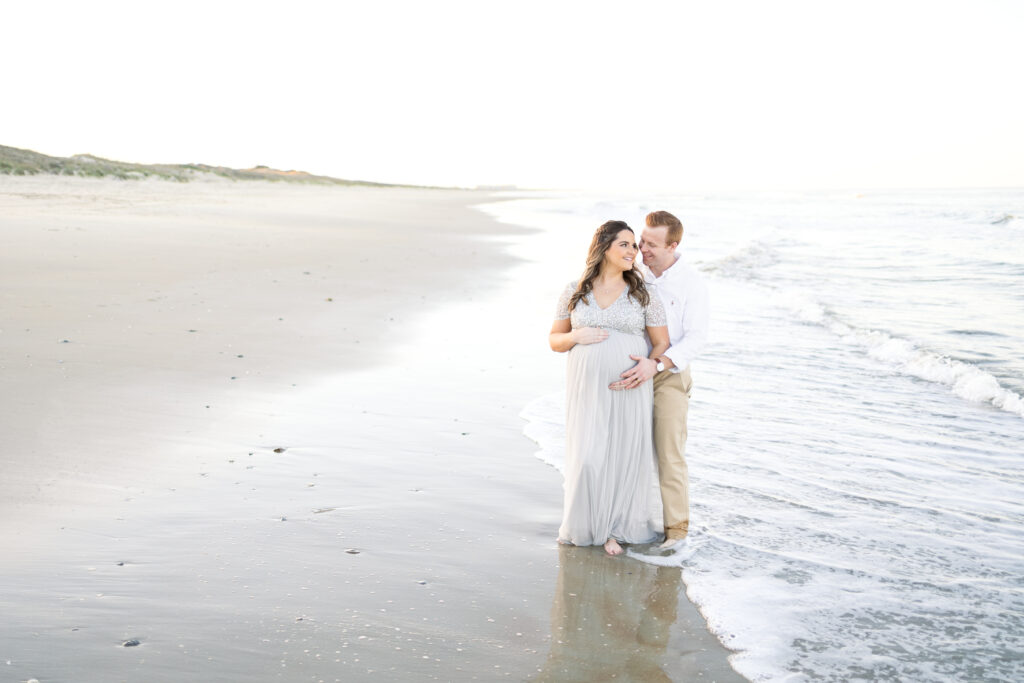 Expectant Mother and Father hugging on the beach during their Virginia Beach maternity portraits with maternity and family photographer Chelsey Kraus