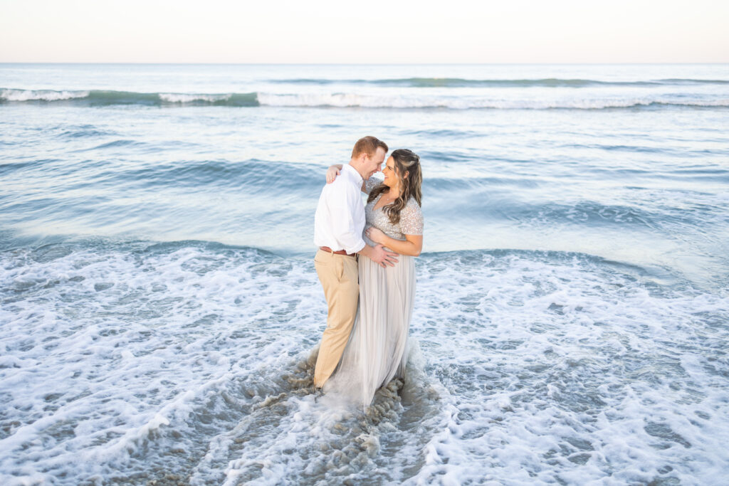 couples snuggling and holding onto pregnant mom's bump during sunset photos on virginia beach