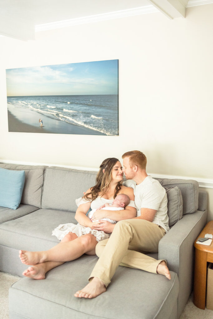 lifestyle family photo at home in virginia beach, mom and dad holding baby girl and going in for a kiss while sitting on the couch underneath a large printed canvas photo from their engagement photos on the beach