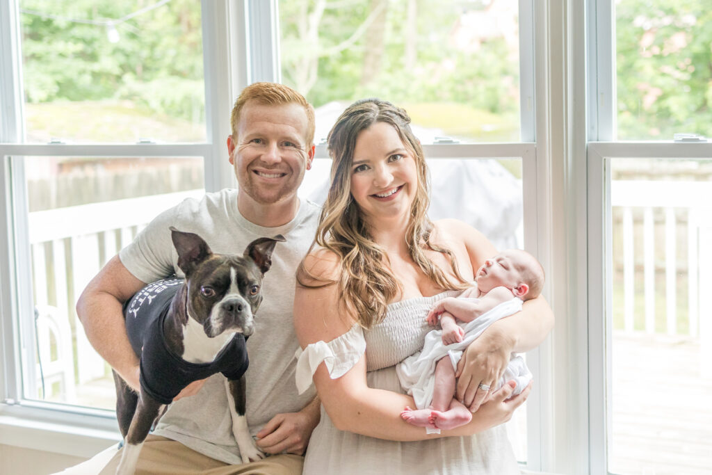 family of 3 posing on bench in home during lifestyle newborn photos in virginia beach with pet dog 
