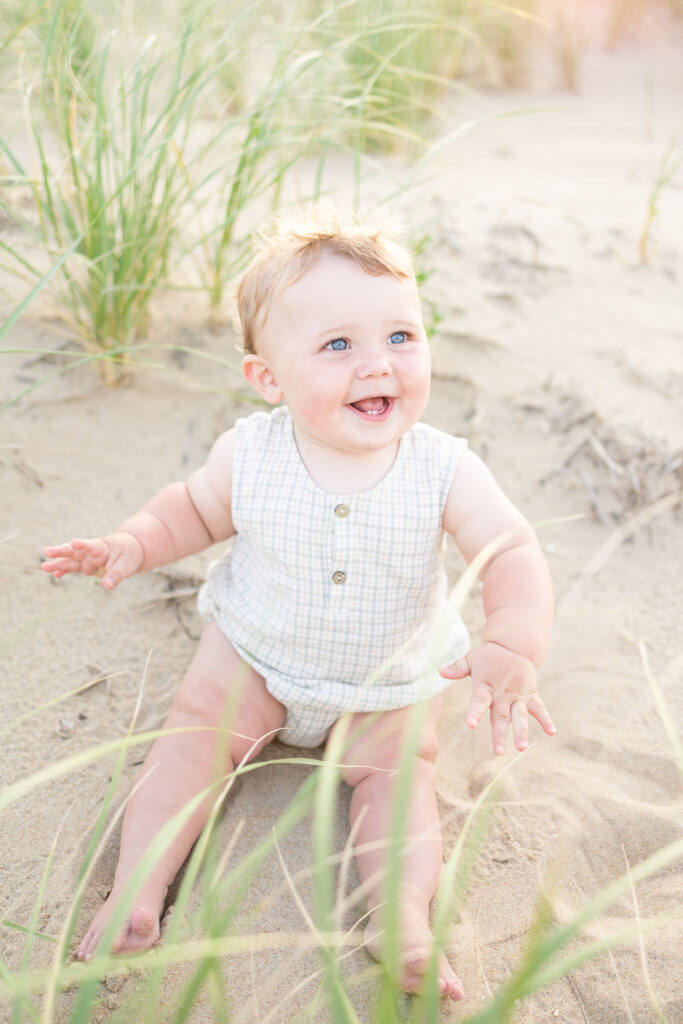 one year old baby boy sitting in the sand dunes on the beach during family photos in virginia beach