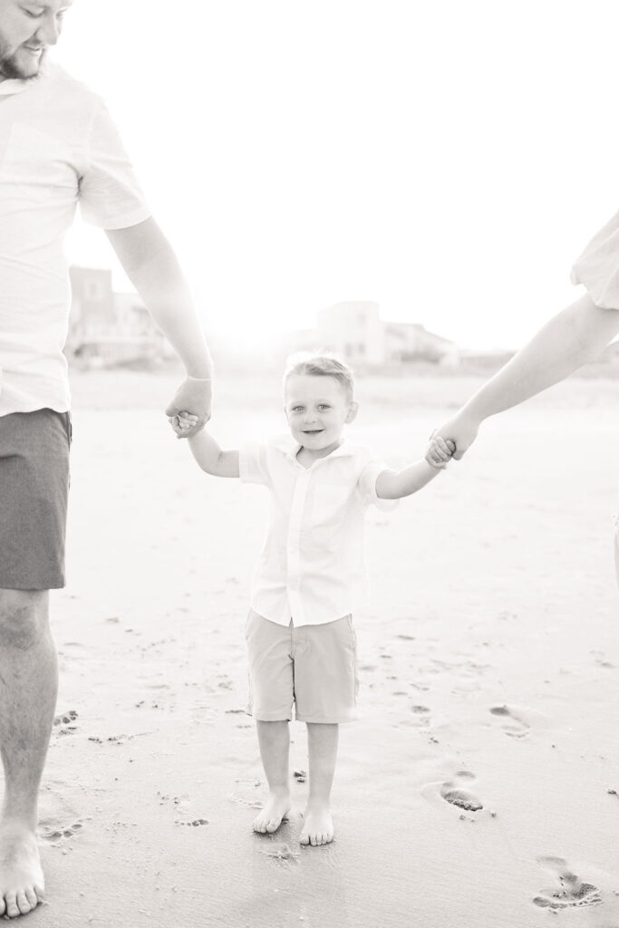 toddler boy smiling while holding hands and walking with his parents during virginia beach family photos 