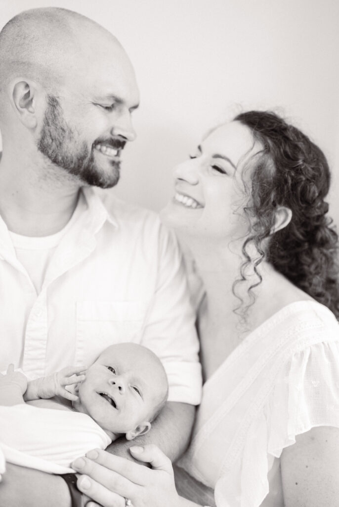 mom and dad holding baby boy during their newborn photo session and smiling at one another 