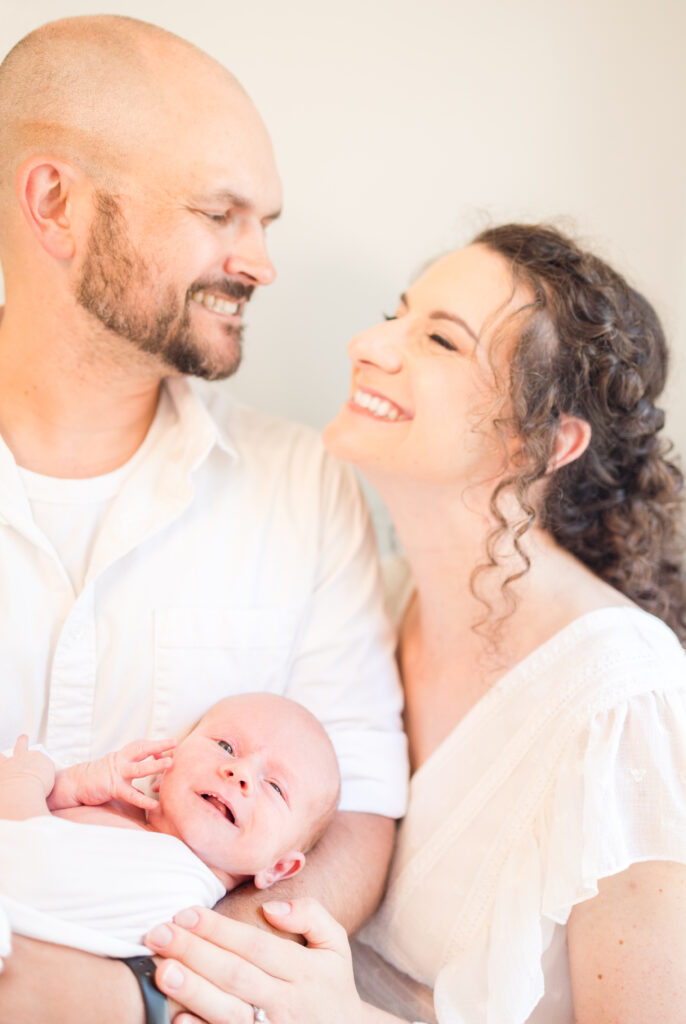 mom and dad smiling at each other during at home newborn photos while holding their baby boy