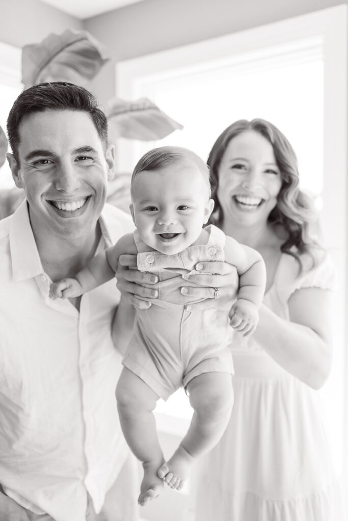 mom holding baby close to camera while she, baby and dad smile during their family photos at home. 