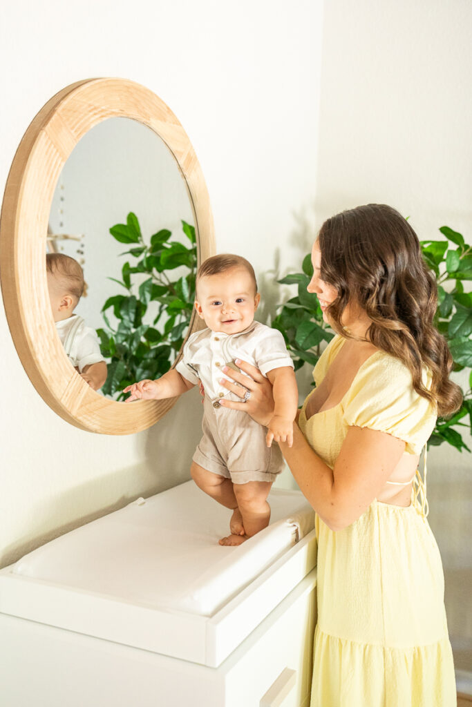 adorable 7 month old baby boy stands on his dresser as mom smiles down at him during their family photos in their home in north carolina
