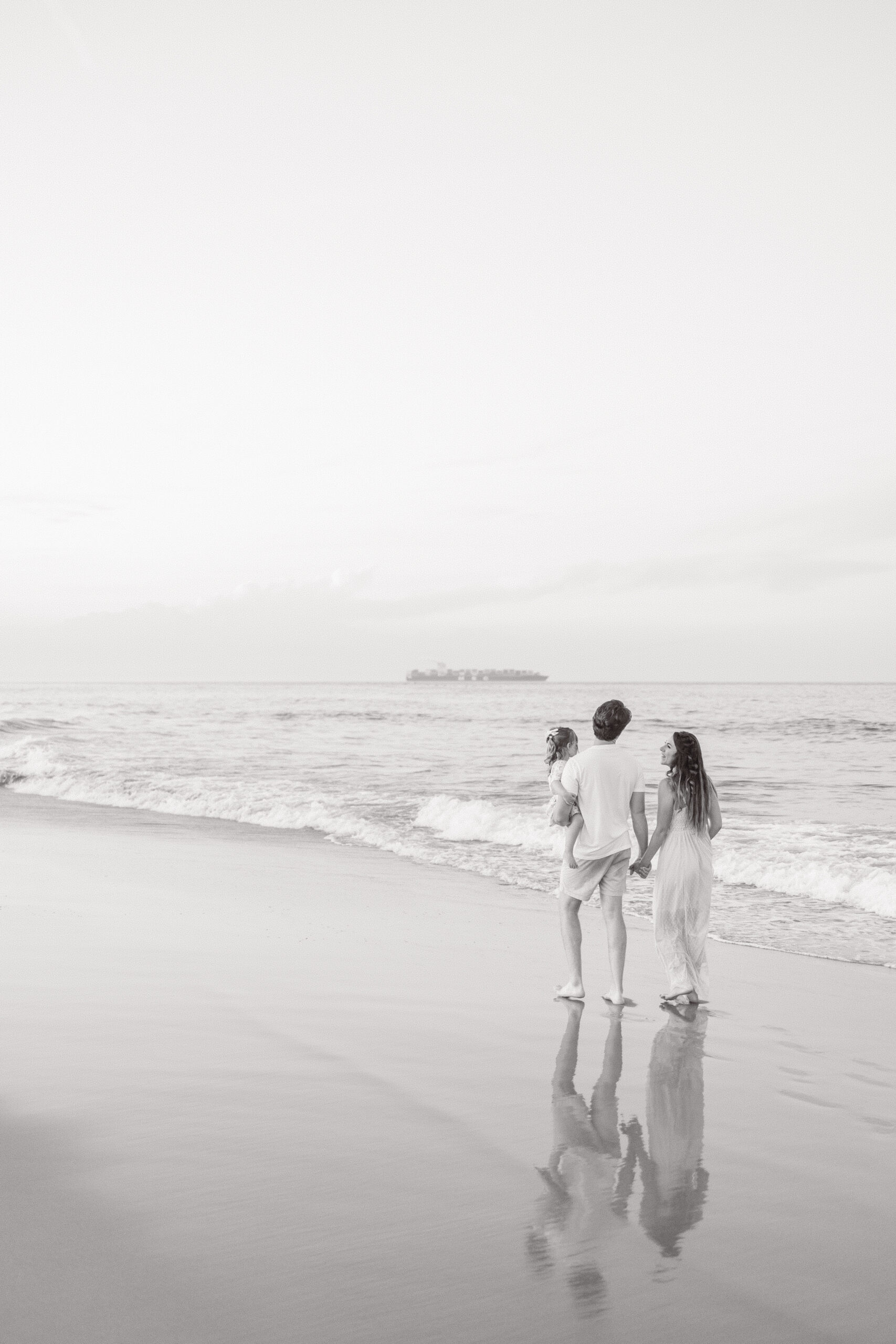 family of 3 walking hand in hand on the edge of the water during sunset family beach photos in virginia beach. 