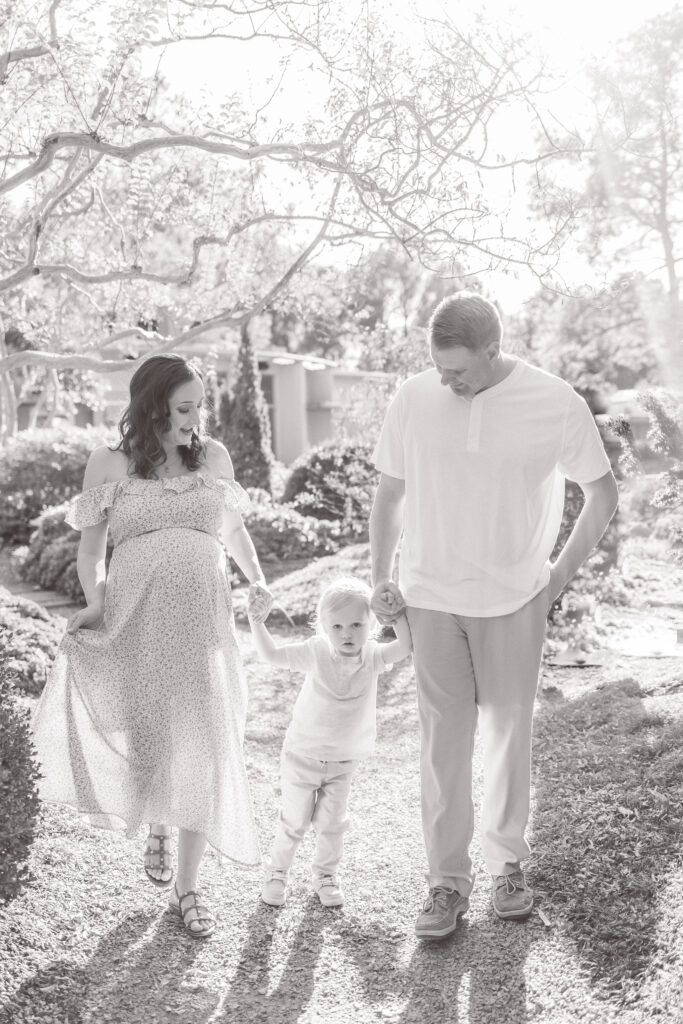 pregnant mom and dad walking hand in hand with toddler boy smiling during maternity photos in norfolk, virginia