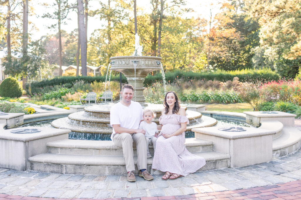 mom, dad and toddler son sitting at a fountain in a botanical gardens during maternity photos in norfolk, virginia, usa 