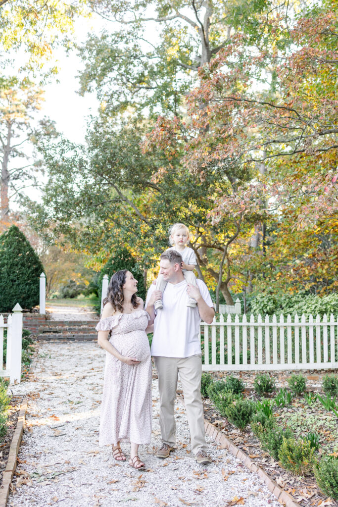 beautiful, candid image of pregnant mother smiling up at her toddler son on his dad's shoulders during maternity photos at the norfolk botanical gardens 