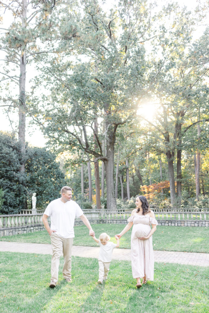 pregnant mom walking hand in hand with toddler son, all are smiling during maternity photos at the norfolk botanical gardens in virginia, usa 