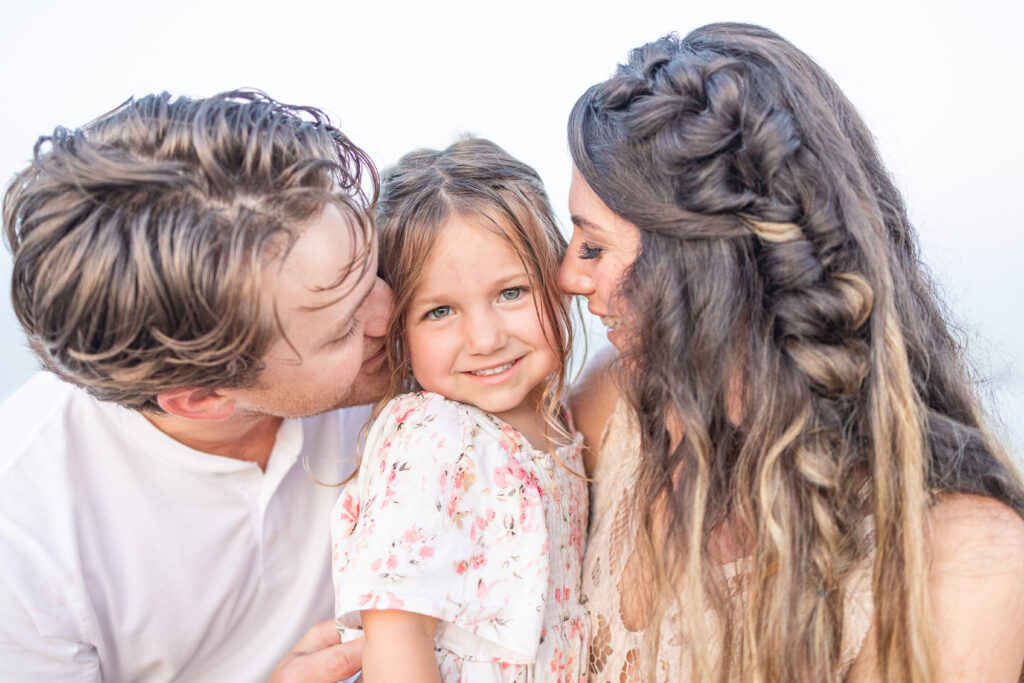 mom and dad snuggling in to kiss baby girl's cheeks during beach photos in virginia beach